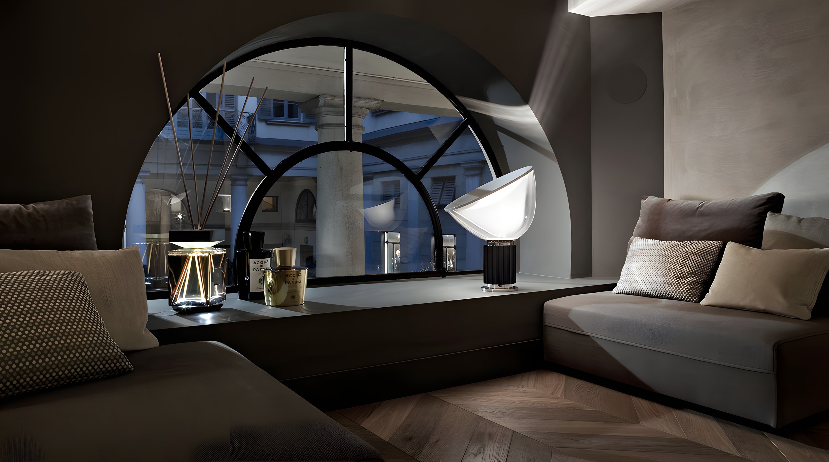 Night-lit interior, Taccia lamp and a diffuser lamp set on a banquette beneath a large arched window, chocolate brown palette and chevron wood floor