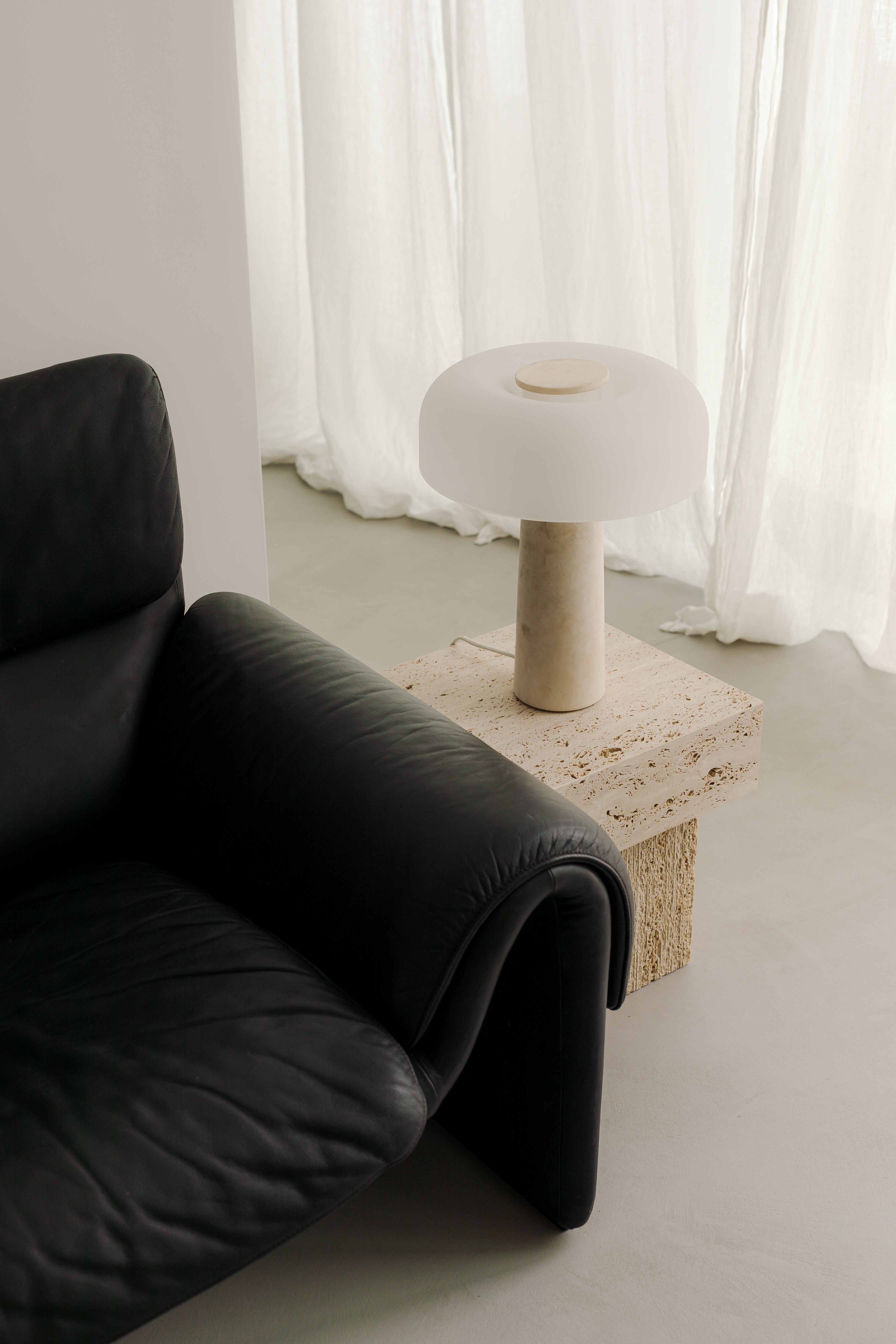 Table d'appoint en travertin aux cavités ouvertes, lampe à pied de pierre claire, fauteuil en cuir noir et rideaux de lin